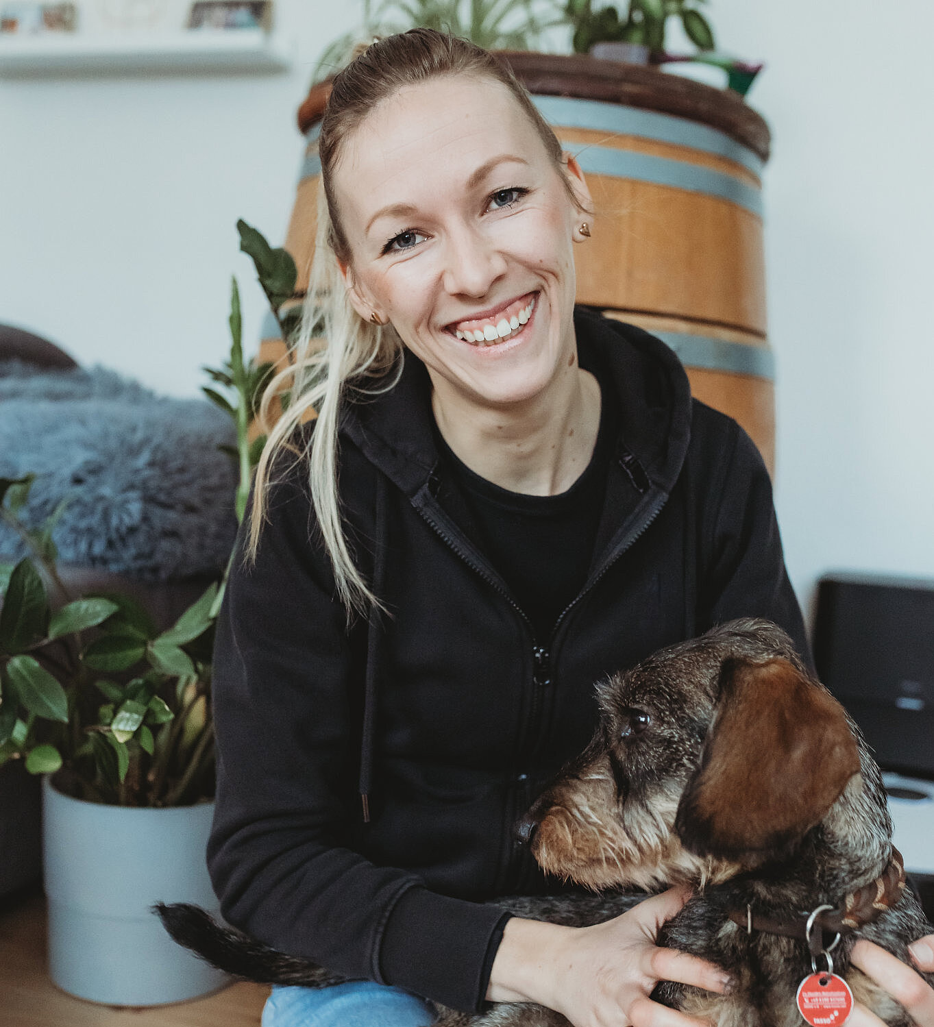 Anna-Lena Heitkötter, mobile Tierphysiotherapeutin, sitzt lächelnd mit einem kleinen Hund auf dem Boden in wohnlicher Umgebung.